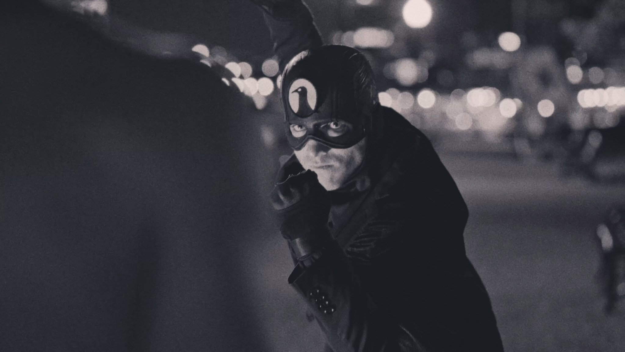 The Raven, masked and in costume, staring toward the camera on a Baltimore street at night.