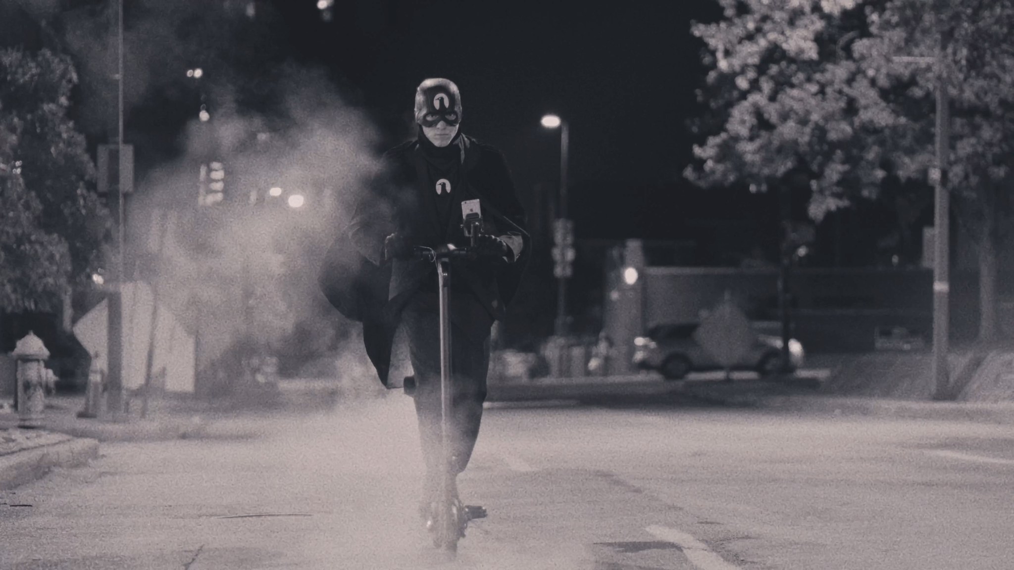 The Raven riding a scooter down a Baltimore street at night through a cloud of smoke.