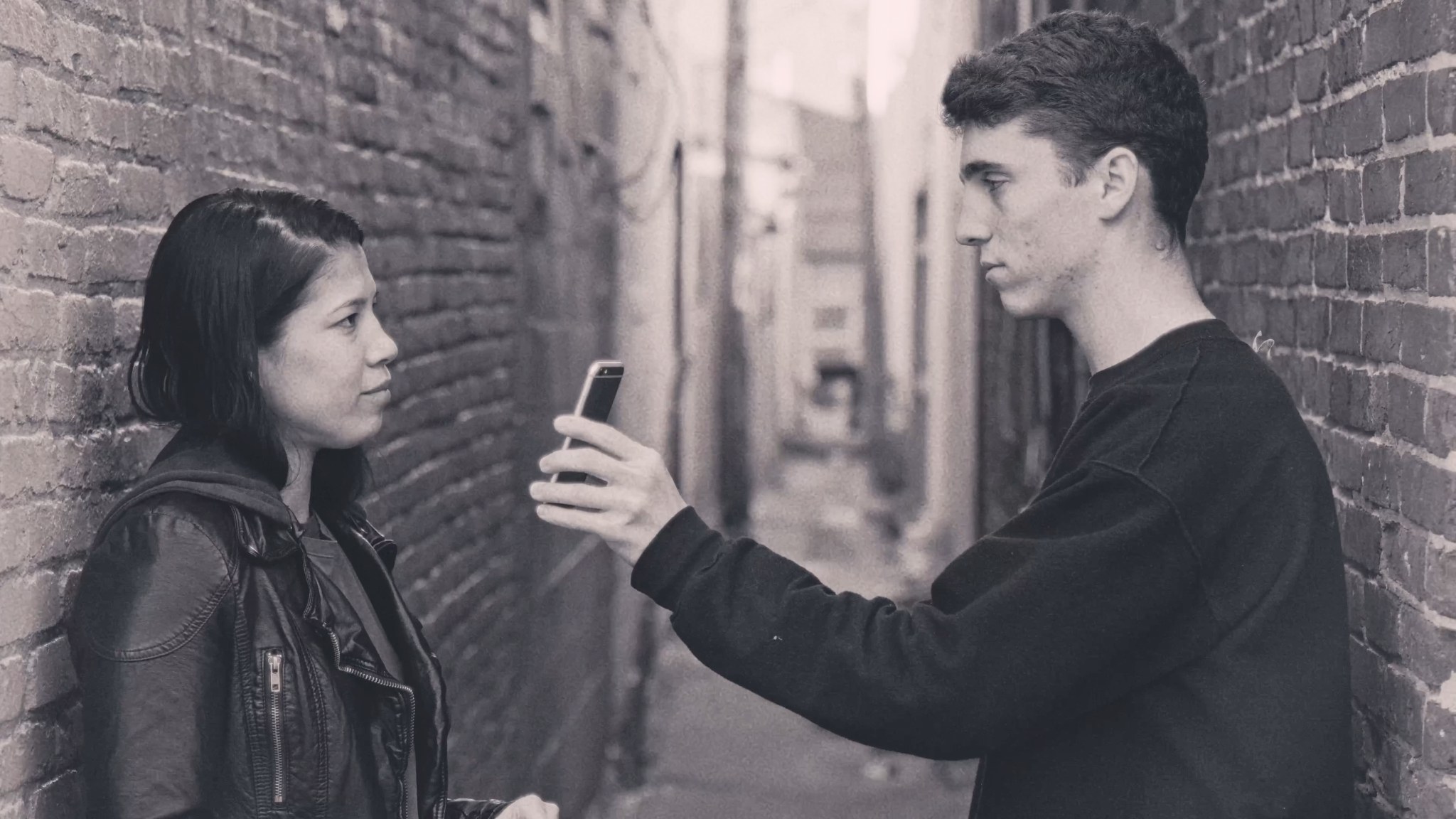 A woman and a man facing one another in a narrow brick alley while he holds up a phone.